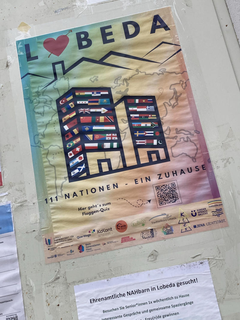 Plakat mit dem Text "Lobeda - 111 Nationen - ein zuhause", zwei Häusern mit
vielen Nationalflaggen und unten Logos der unterstützenden
Organisationen