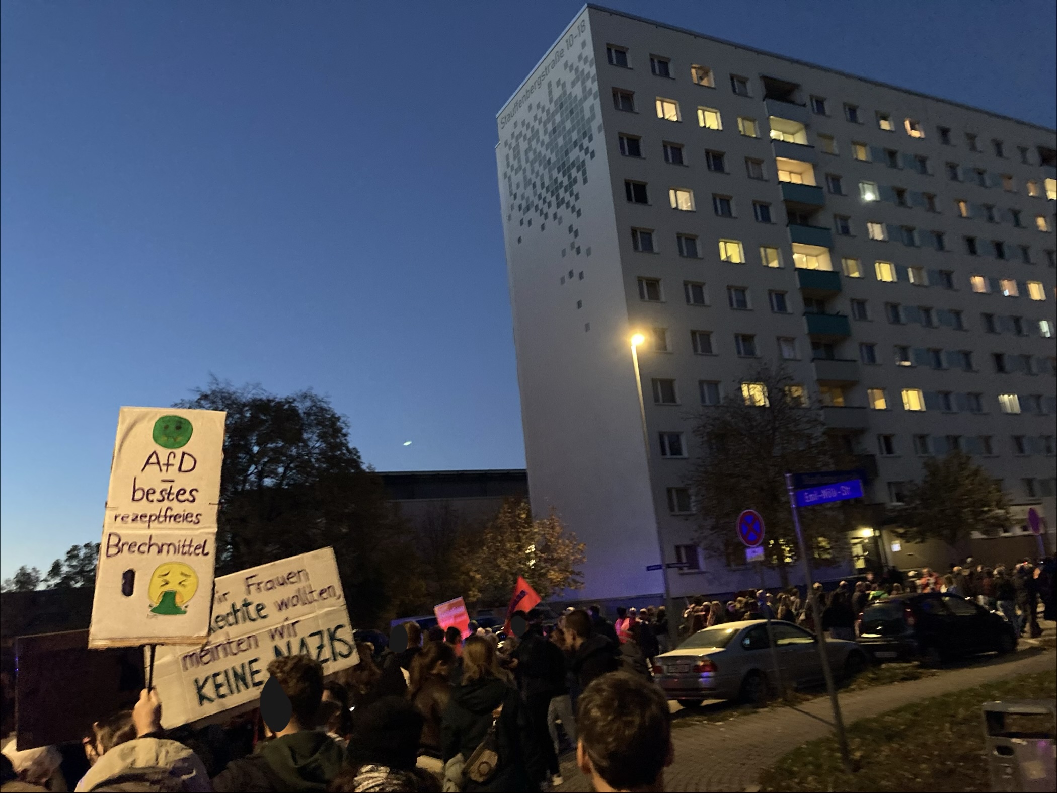 Viele Menschen sind von hinten auf einer Demo zu sehen, die vor einem Plattenbau läuft. Eine Person trägt ein Schild mit dem Text 'AfD - bestes rezeptfreies Brechmittel'