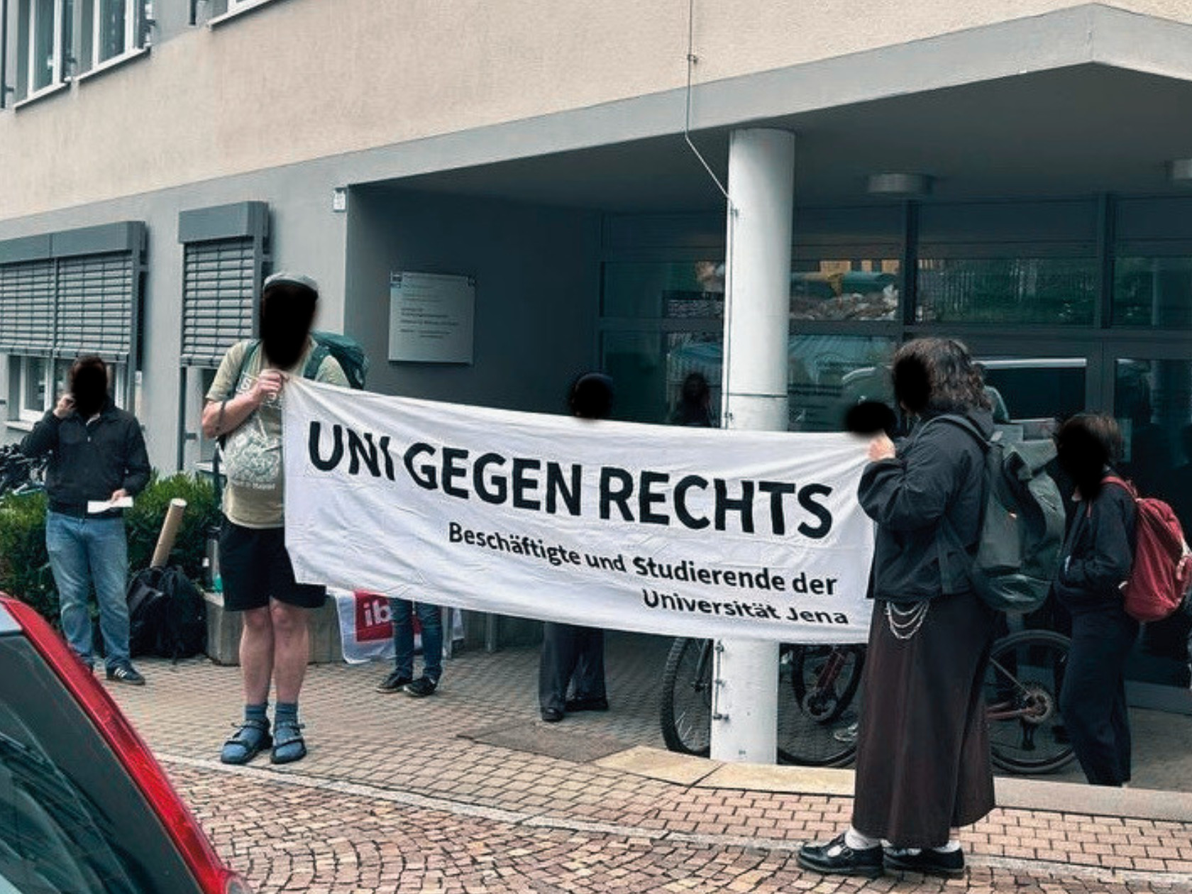 Zwei Personen halten ein Uni gegen Rechts Transparent vor dem Gebäude der Erziehungswissenschaften. Um sie herum sind weitere Personen zu sehen.